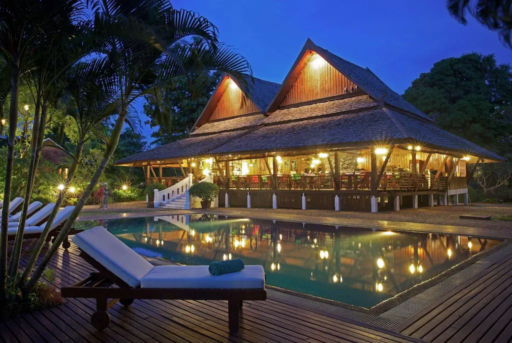 Illuminated wooden pavilion of La Folie Lodge reflecting in a pool at dusk