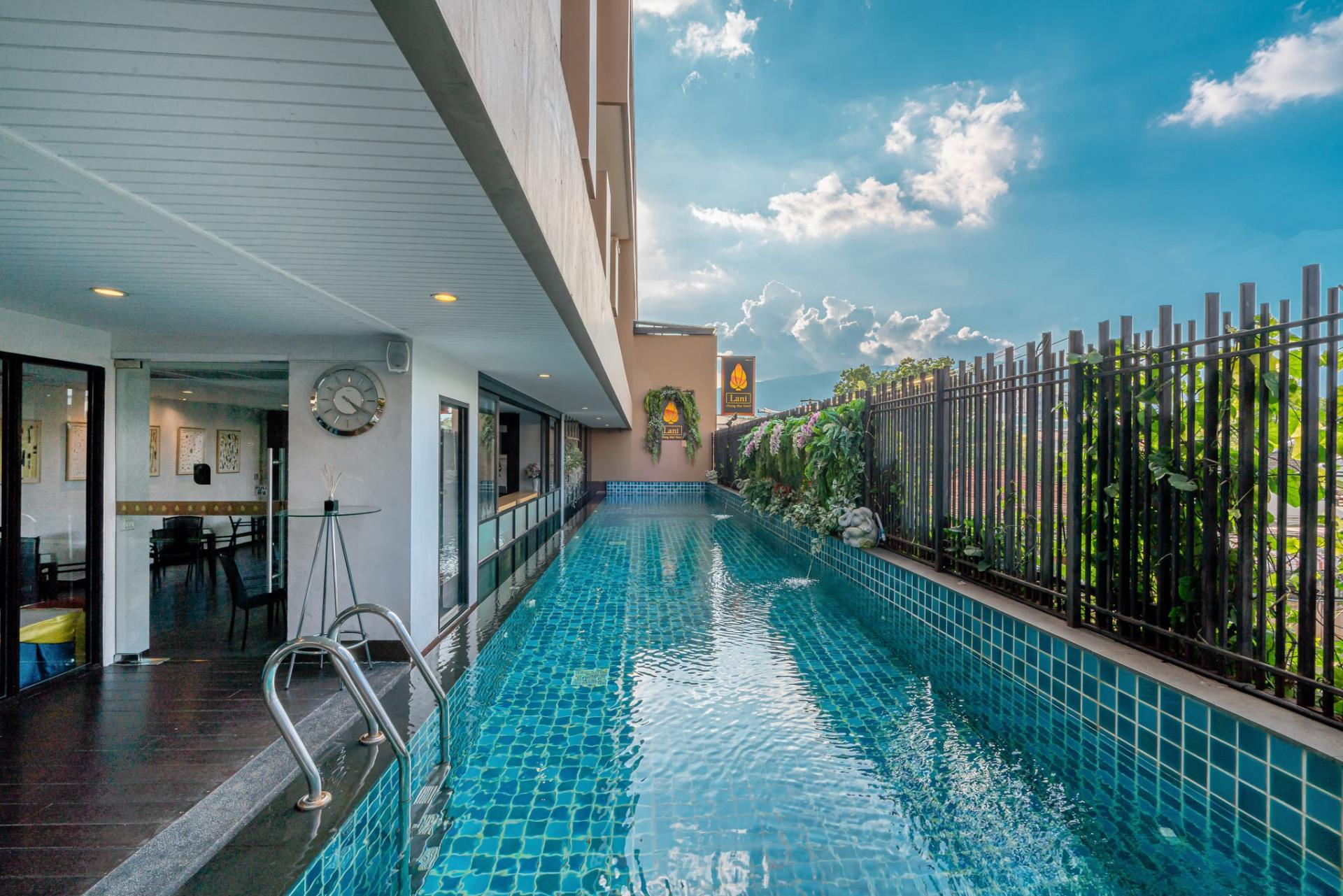 Long narrow swimming pool next to building and garden fence with plants under blue sky