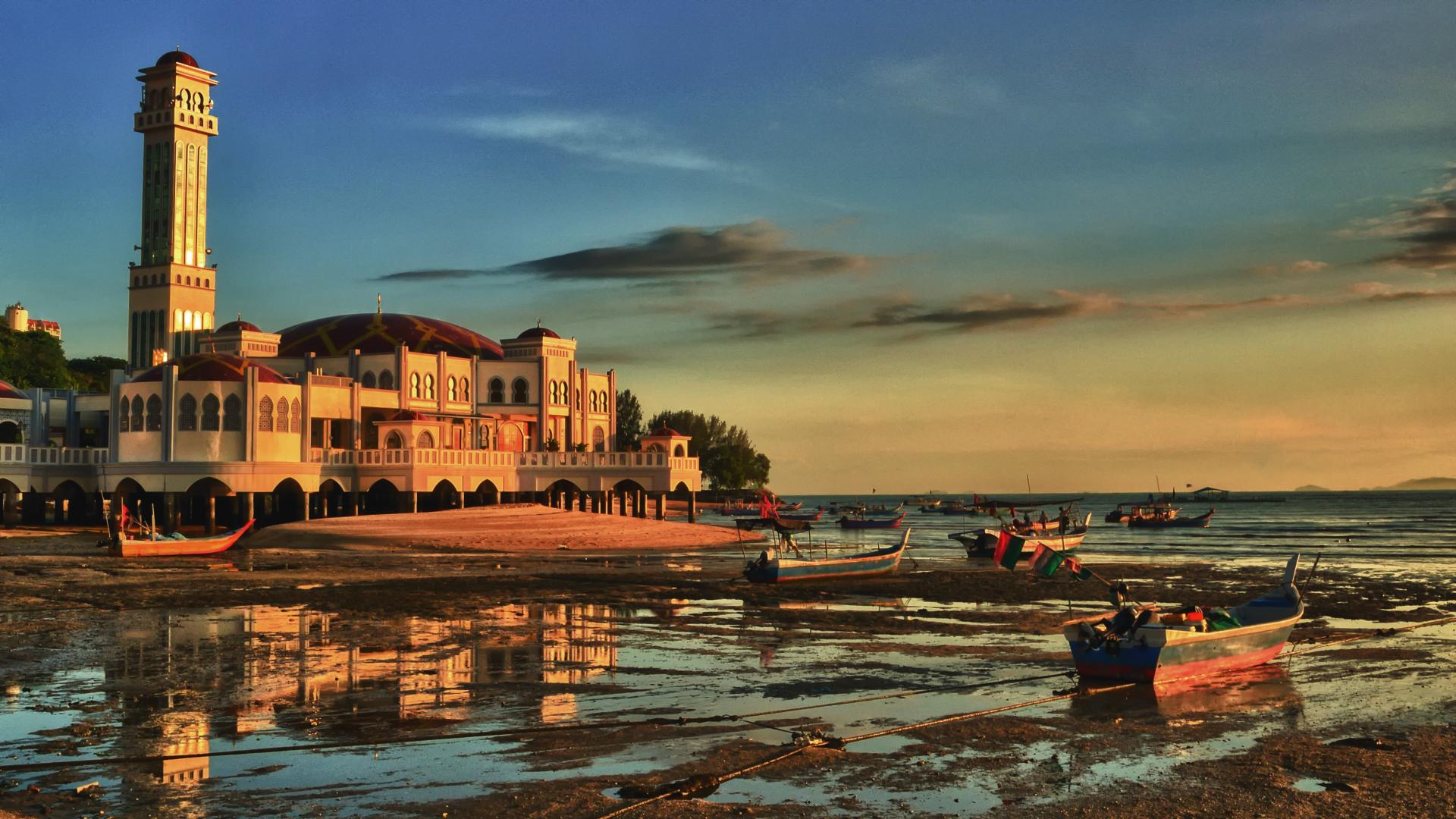 Coastal mosque lit by sunset with boats resting on shore at low tide