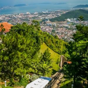 Red and white funicular train ascending green hill with city and sea in background