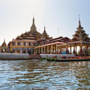 Burmese temple with golden spires by lake and docked colorful longboat