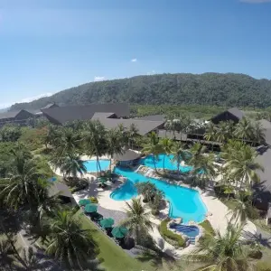 Aerial view of a tropical resort pool surrounded by palm trees and buildings with mountains in background.