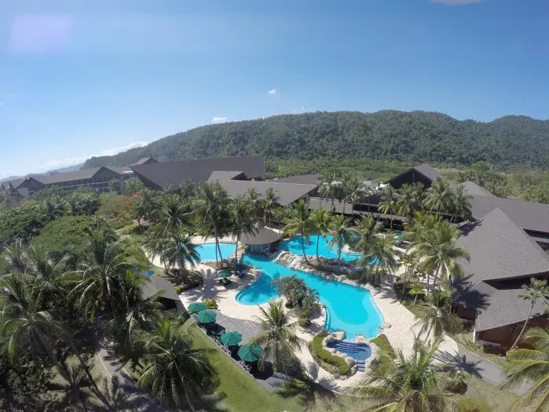 Aerial view of a tropical resort pool surrounded by palm trees and buildings with mountains in background.