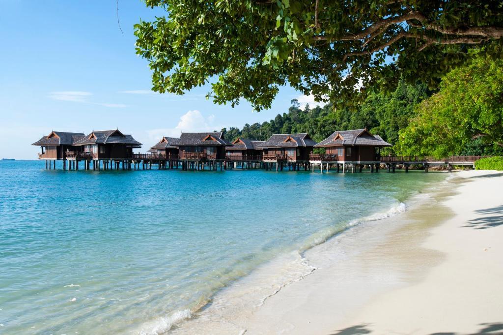 Wooden overwater villas on stilts by tropical beach with calm blue sea and green trees.