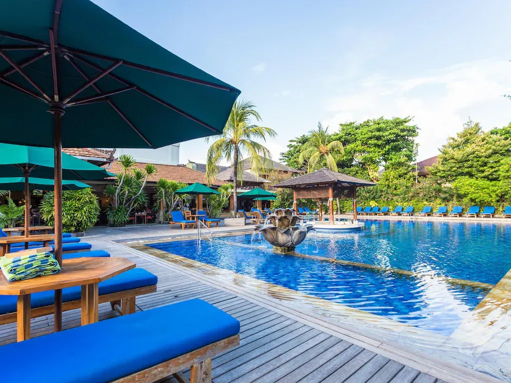 Hotel outdoor pool with blue loungers, green umbrellas, fountain, and palm trees