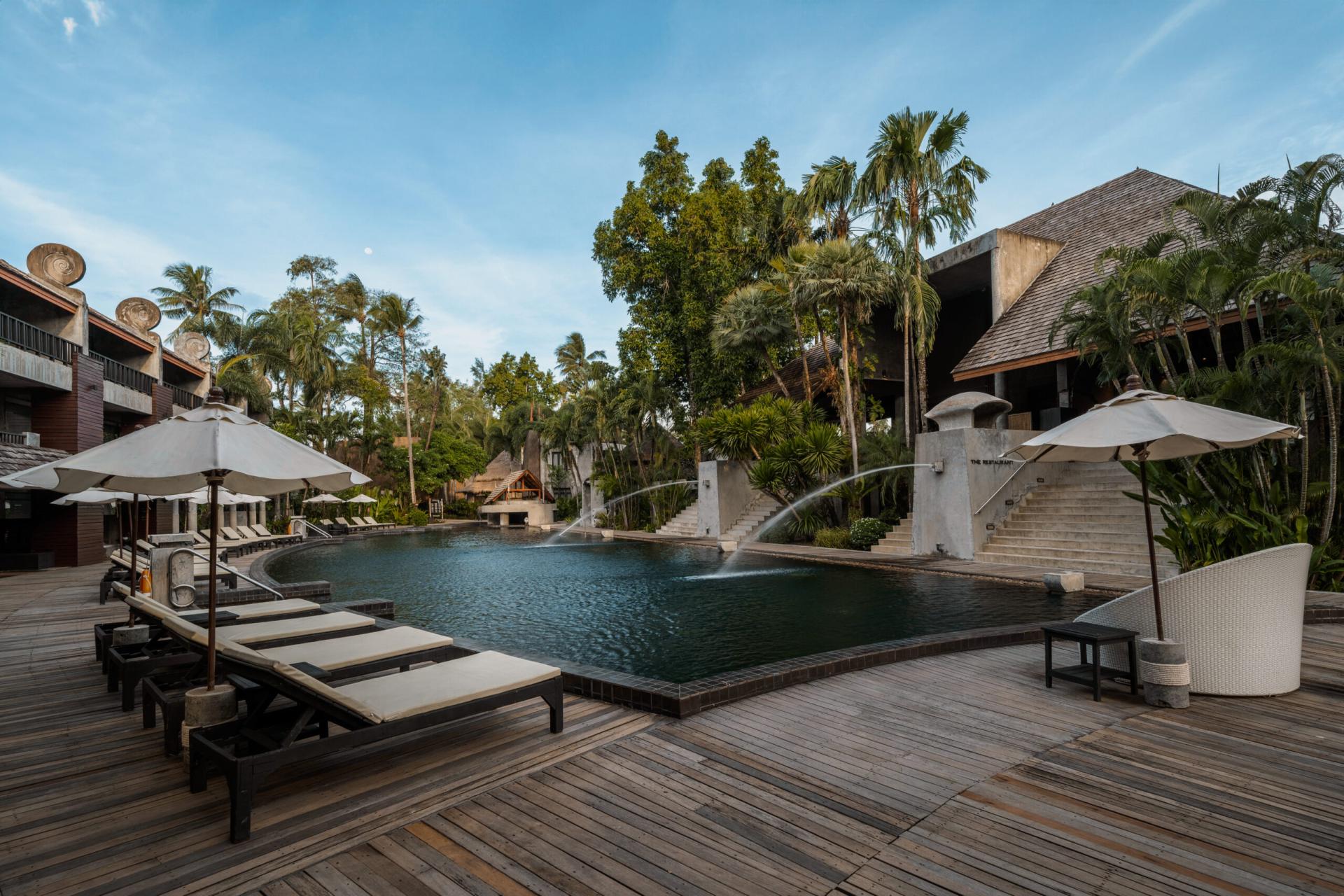 Resort pool with sun loungers, umbrellas, water fountains, palm trees, and wooden deck