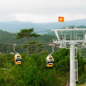 Cable cars moving over green pine forest with hills in background and Vietnam flag on tower
