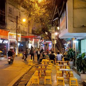 Night street with outdoor yellow stools, people dining, walking, and a motorbike passing