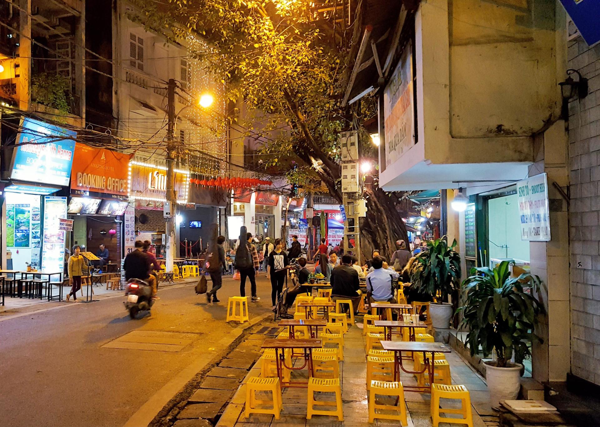 Night street with outdoor yellow stools, people dining, walking, and a motorbike passing