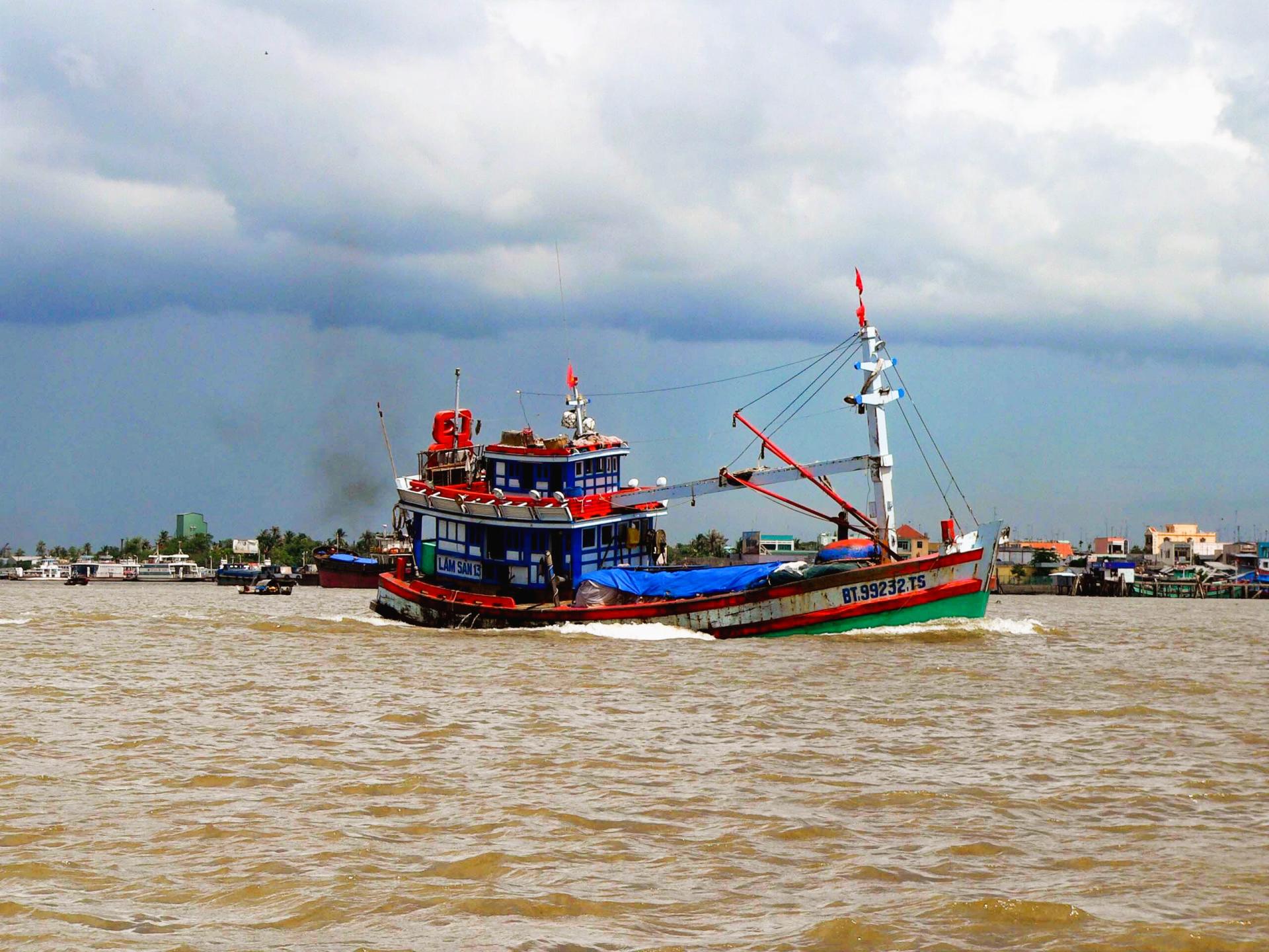 Colorful traditional wooden boat moving on muddy river near buildings under cloudy sky
