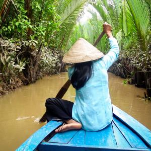 Woman in conical hat paddling blue boat through palm-lined muddy river canal