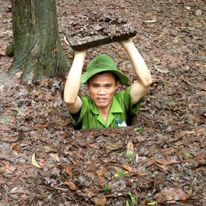 Man lifting leaf-covered trapdoor to exit camouflaged tunnel in forest