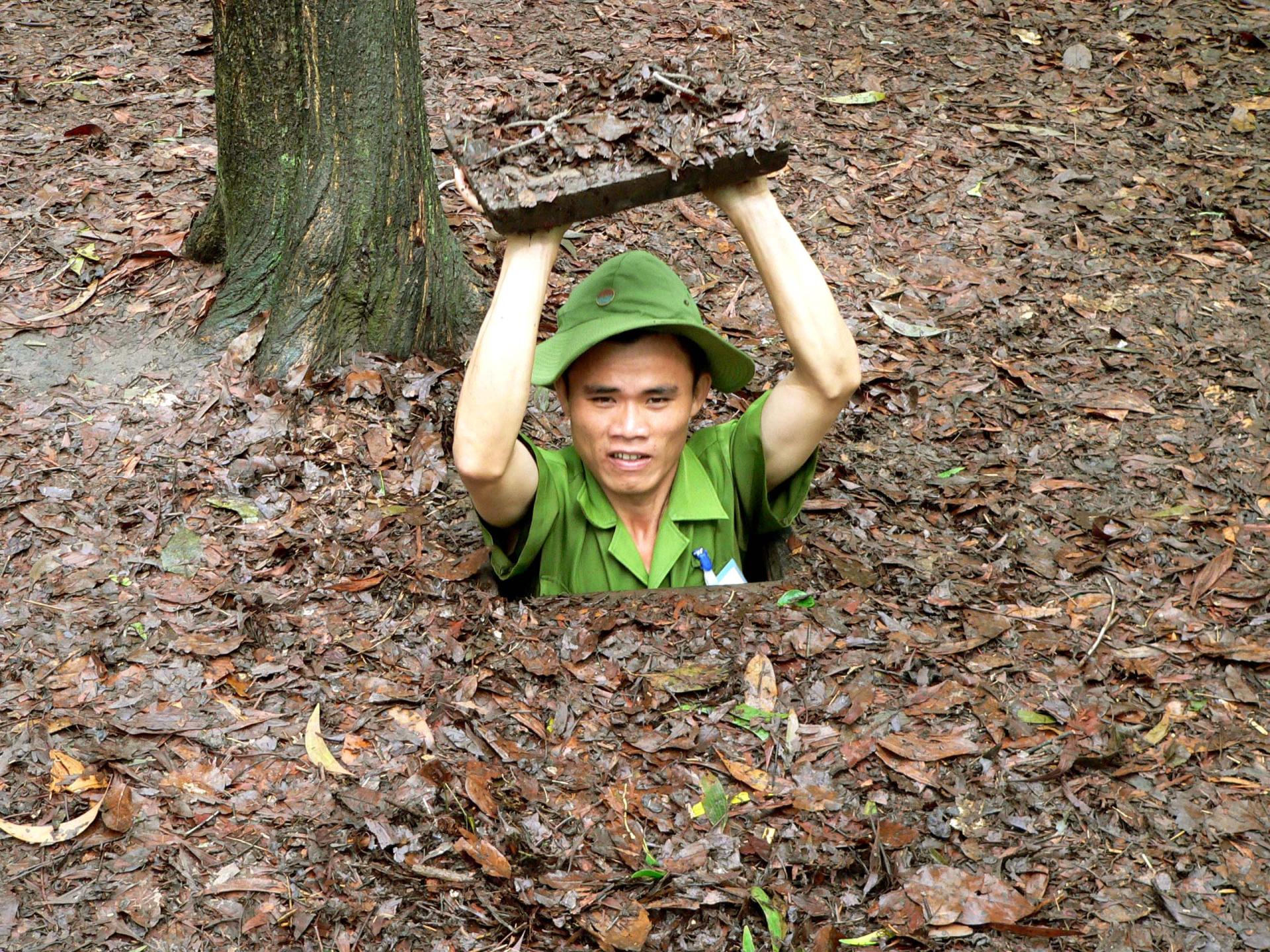 Man lifting leaf-covered trapdoor to exit camouflaged tunnel in forest