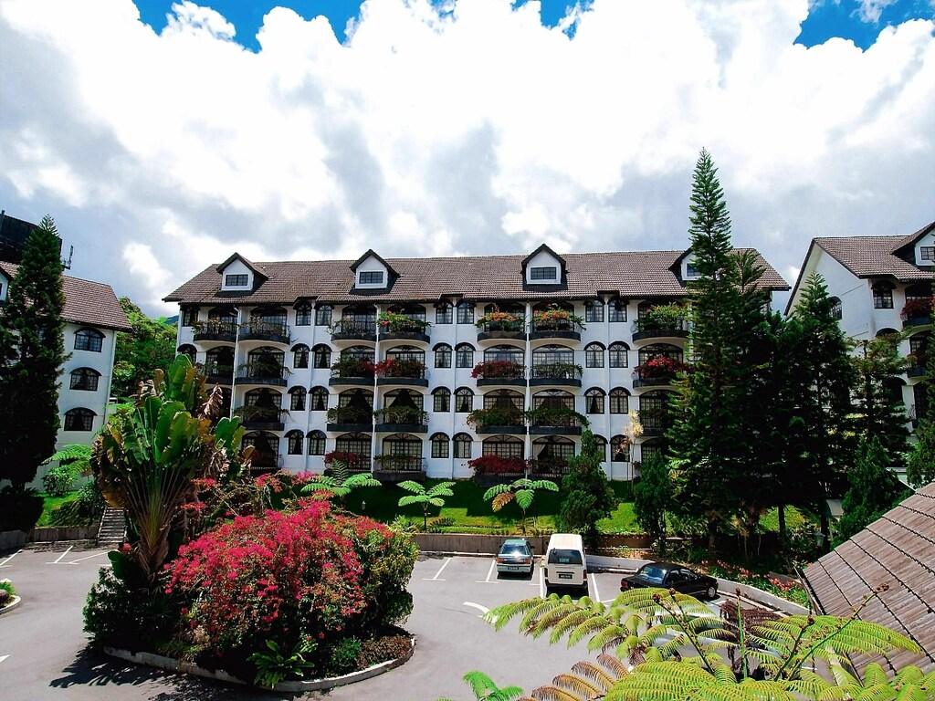 White hotel building with balconies, garden, parked cars, and cloudy sky