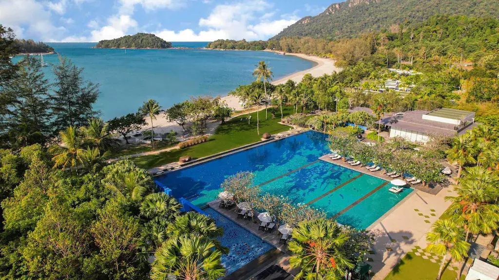 A large outdoor swimming pool at a beach resort with tropical trees and a sandy beach nearby