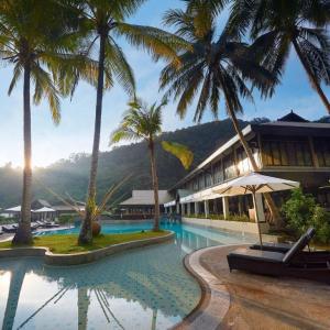 Resort pool with palm trees, sun loungers, umbrellas, hotel building, and hills